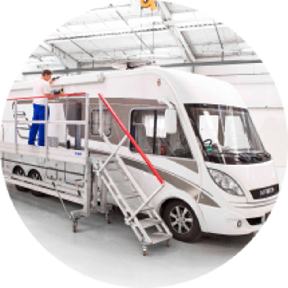 Weißes Wohnmobil in einer Montagehalle mit einer verstellbaren Aluminium-Arbeitsbühne für Reparatur- und Servicearbeiten.
