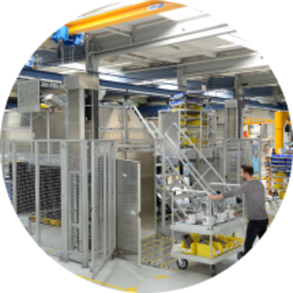 Industrielle Fertigungsanlage mit Schutzgitter-Einhausung, Laufstegen und Mitarbeiter mit Werkzeugwagen in der Produktion.
